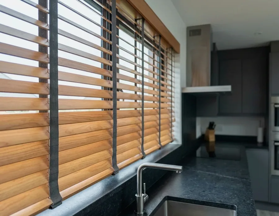Timber venetian blinds on woven ladder tapes across a kitchen window, slats tilted to cut the glare